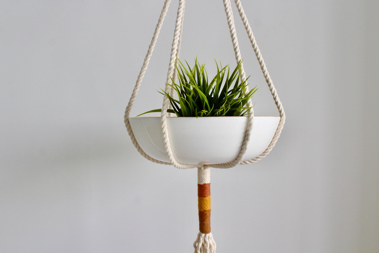 A white ceramic hanging planter suspended by thick cream macrame ropes, holding a small green succulent plant, against a plain light gray background.