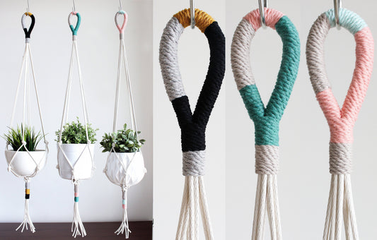 Four macrame plant hangers displayed against a plain light background, with three holding white pots containing green plants and one shown empty. The hangers feature chunky woven rope in black, teal, pink, white, and mustard yellow color blocking.