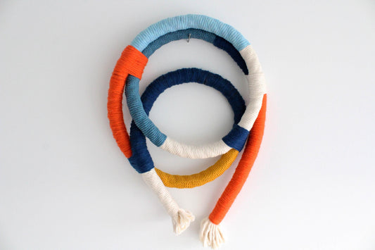 A handmade wall hanging headband made from thick, textured yarn wrapped in horizontal bands of light blue, orange, navy, cream, and mustard yellow, photographed on a clean white background.