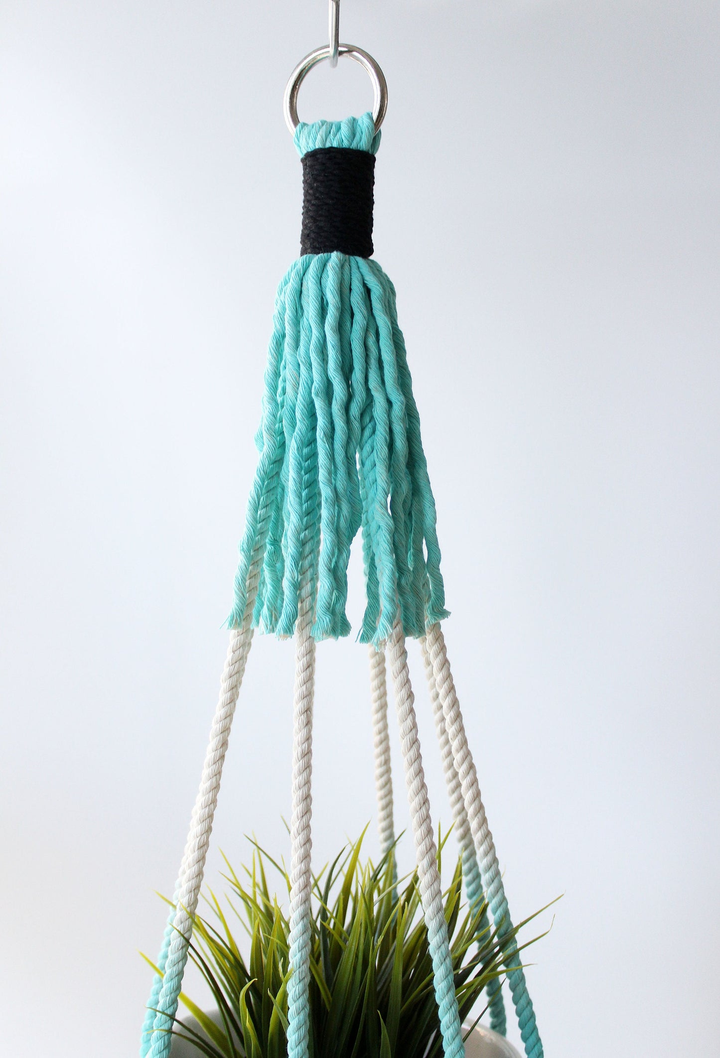 A hanging macrame plant hanger with a turquoise and black woven top, white rope strands, and a silver metal ring, shown holding a green plant against a light gray background.