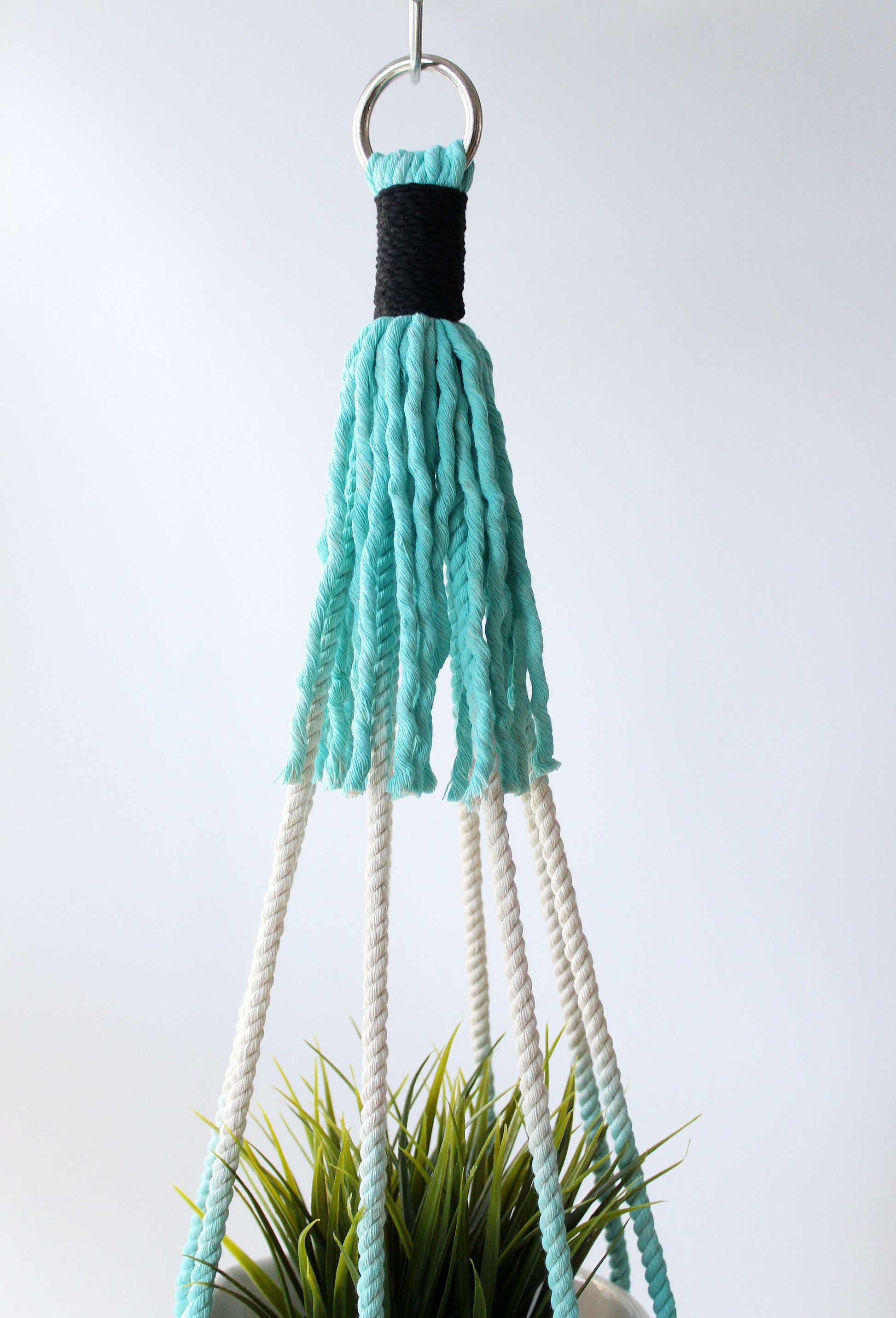 A hanging macrame plant hanger with a turquoise and black woven top, white rope strands, and a silver metal ring, shown holding a green plant against a light gray background.