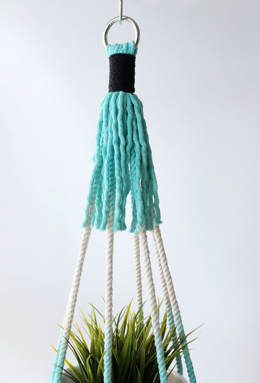 A hanging macrame plant hanger with a turquoise and black woven top, white rope strands, and a silver metal ring, shown holding a green plant against a light gray background.