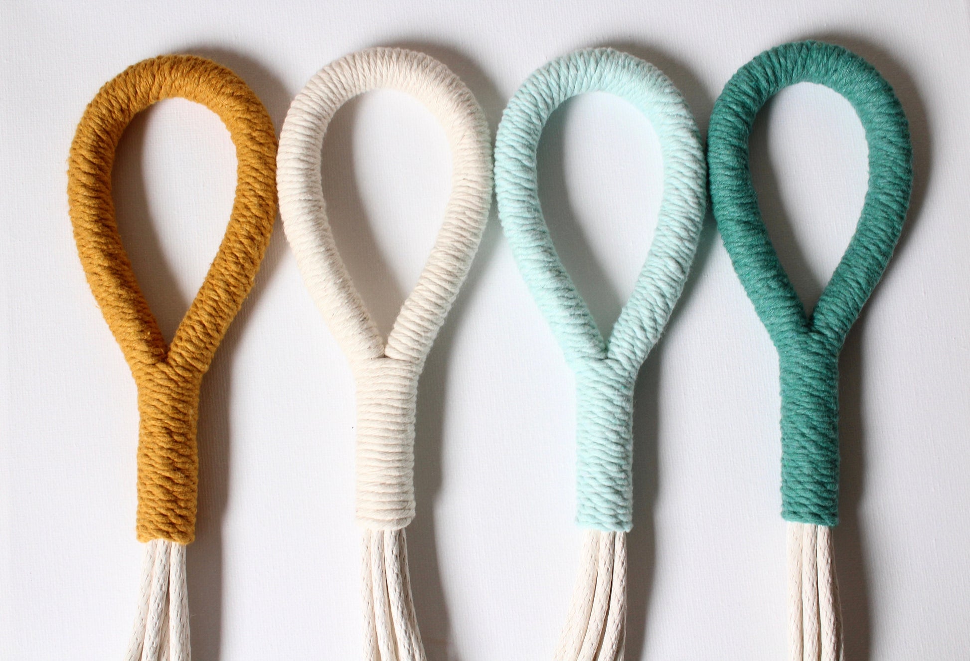 Four rope loop wall hooks in mustard yellow, cream, mint green, and teal, arranged in a row against a plain white background.