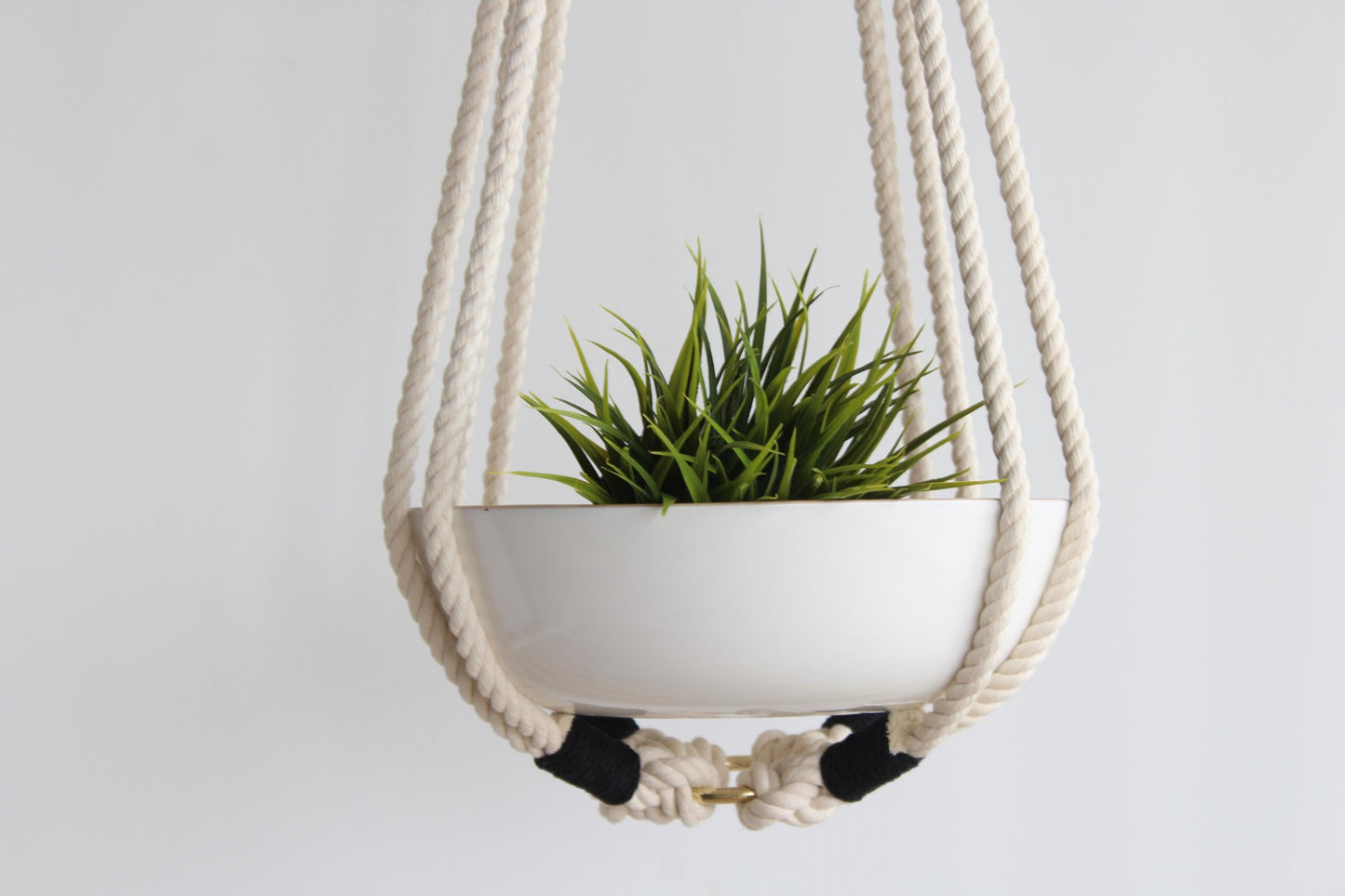 A white ceramic hanging planter suspended in a cream cotton rope macrame hanger, containing a green grass-like plant, set against a plain light gray background.