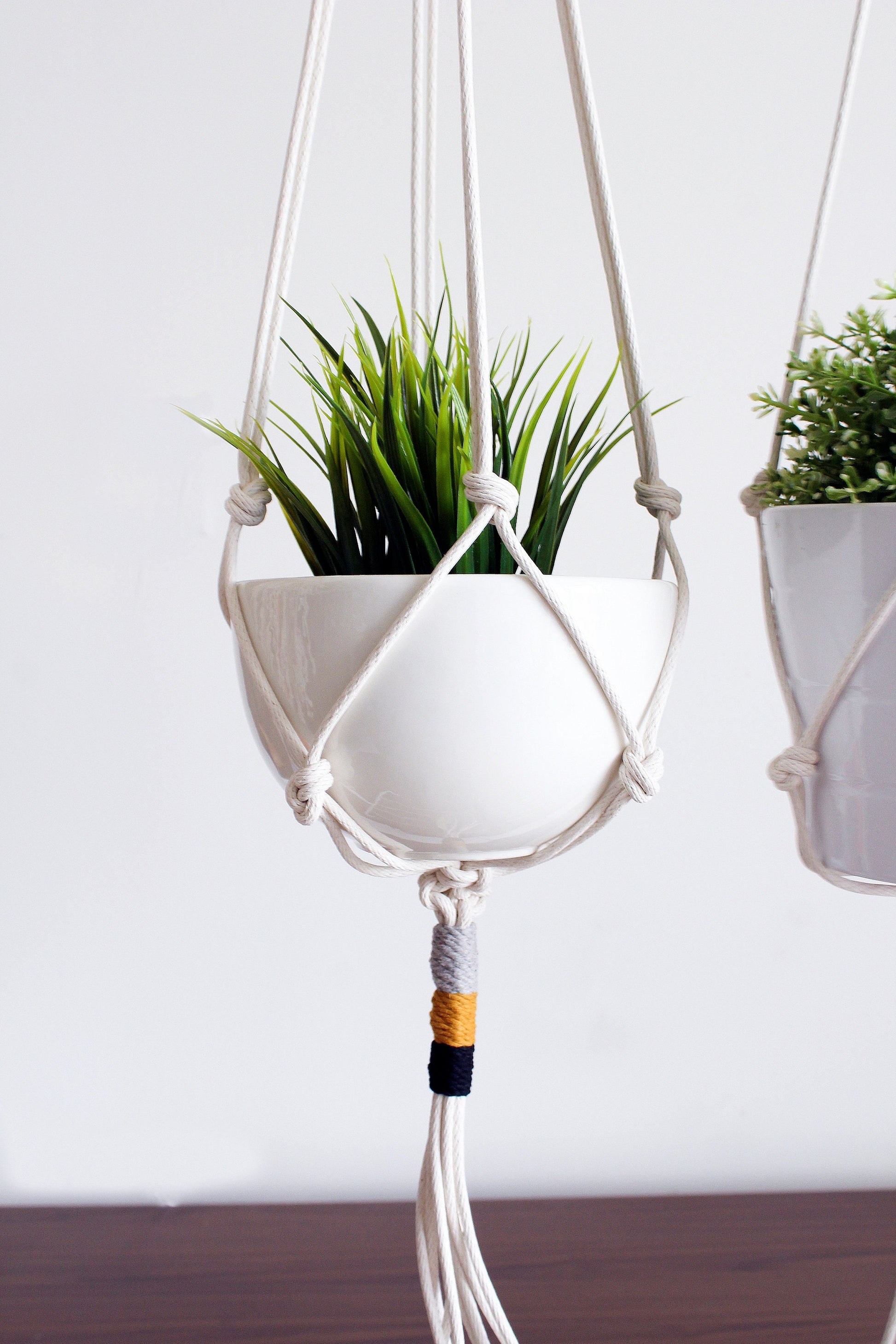 A white ceramic planter in a macrame hanger suspended against a light gray wall, holding a green artificial plant. Another matching planter is partially visible on the right.