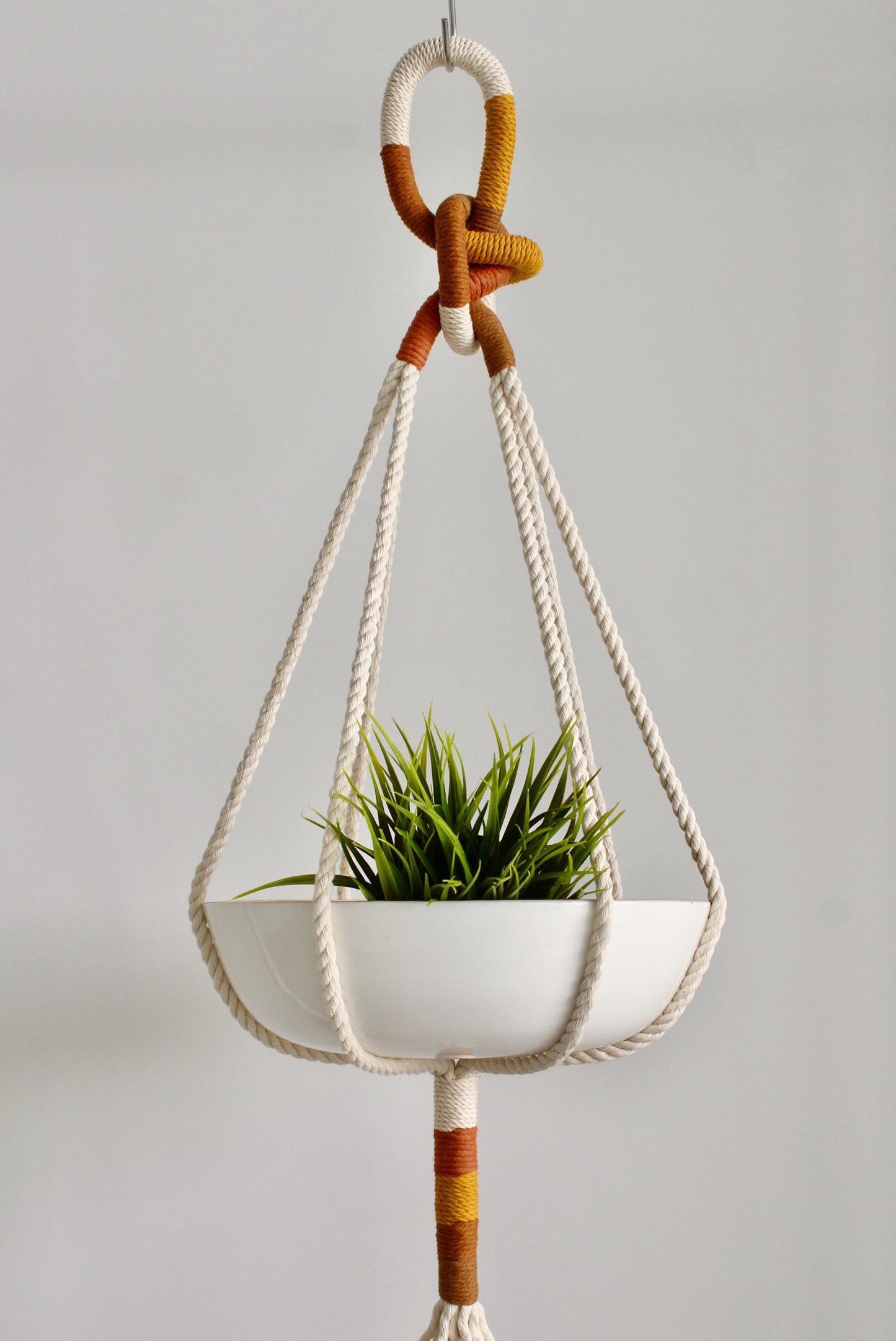 A white ceramic hanging planter with a green succulent plant, suspended by a cream rope macrame hanger with orange and brown striped accents, set against a plain light gray background.