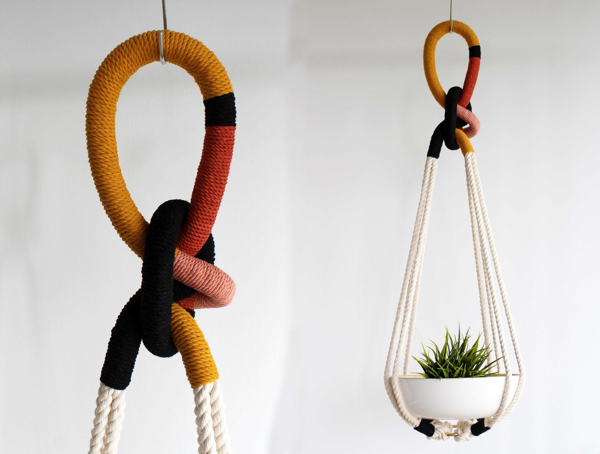 A hanging macrame plant holder with a woven rope loop in mustard yellow, terracotta red, dusty pink, and black, suspending a white planter with a small green succulent, shown against a plain white wall.