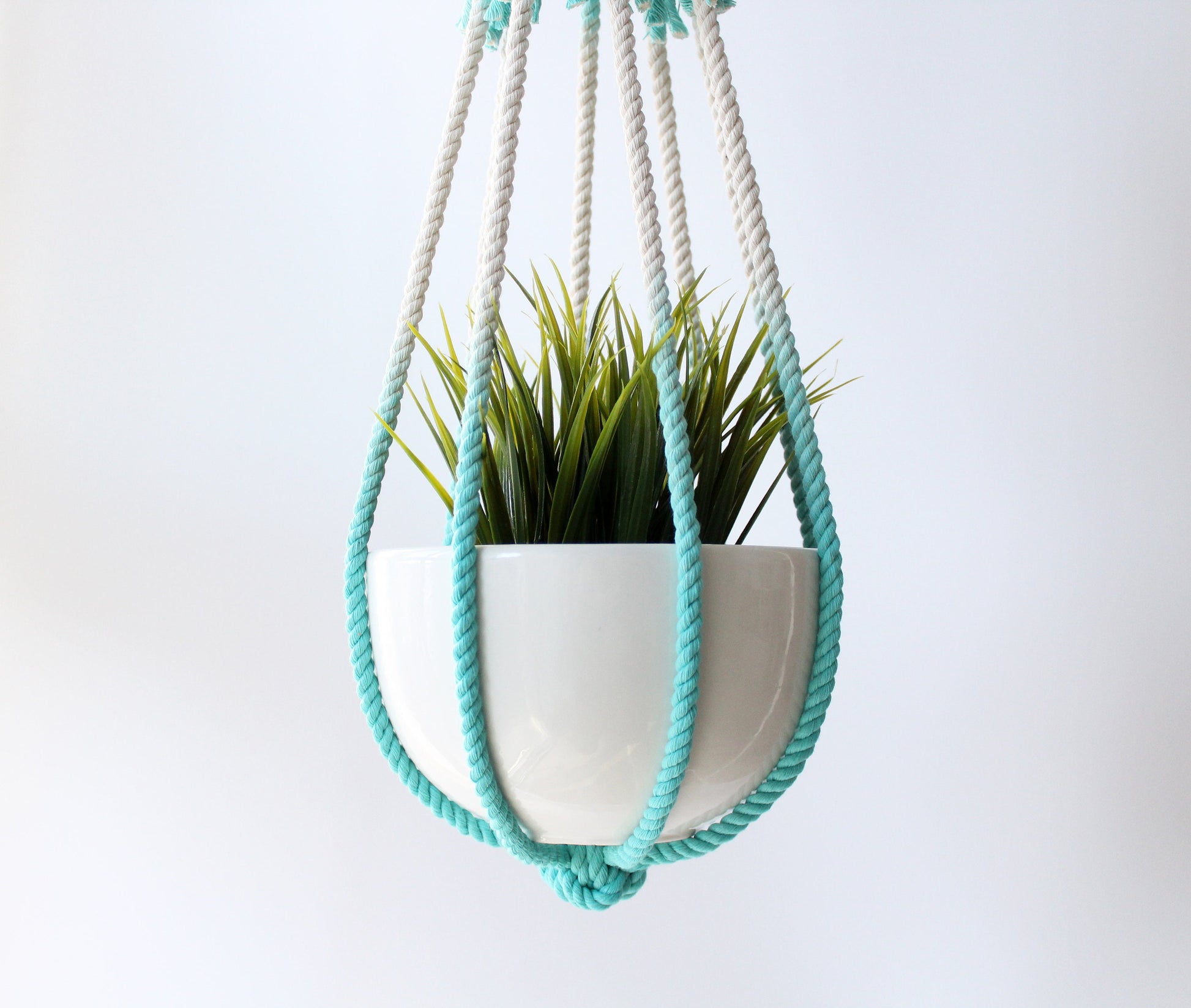 A white ceramic hanging planter with a green artificial plant, suspended in a white and turquoise rope macrame hanger against a plain white background.