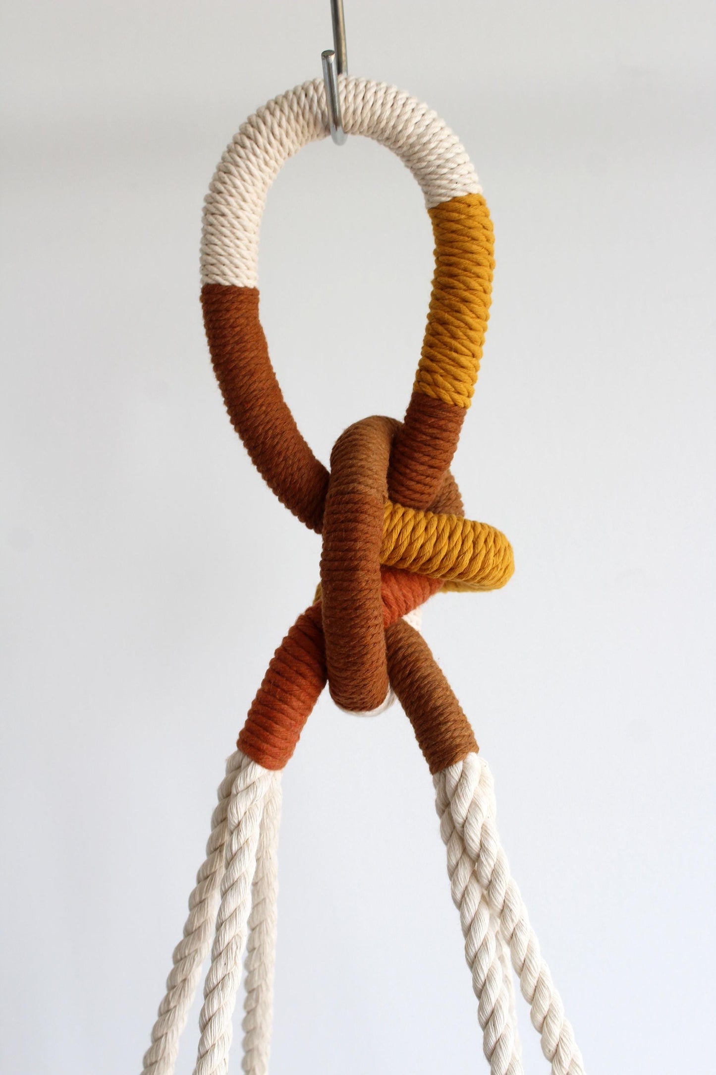 A close-up of a decorative rope knot hanging from a metal hook against a plain light background. The rope features a color-blocked pattern in cream, terracotta, mustard yellow, and orange.