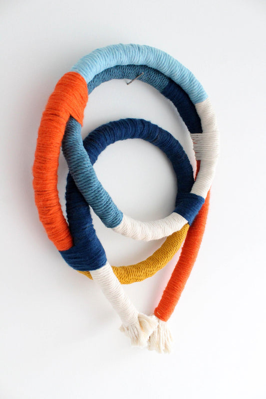 A bohemian-style woven wall hanging made from three interlocking circular hoops wrapped in textured yarn. The hoops feature stripes of orange, light blue, dark blue, white, and mustard yellow, with cream-colored tassels at the bottom, photographed on a clean white background.