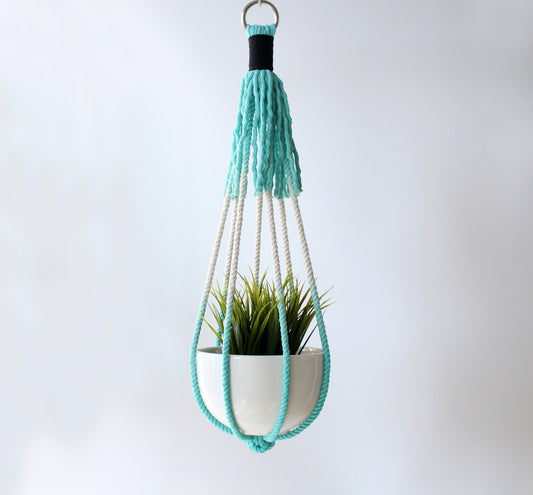 A mint green macrame plant hanger holding a white ceramic pot with a green artificial plant, suspended against a light gray background.
