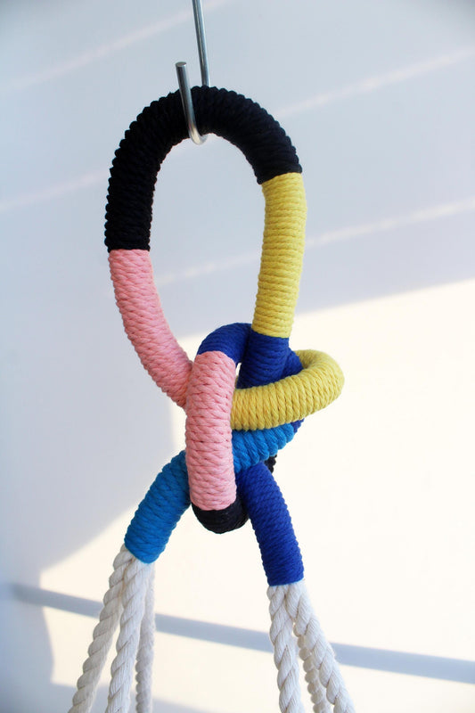A colorful rope climbing knot or hanging loop with black, pink, yellow, and blue textured rope sections, suspended against a bright white background.